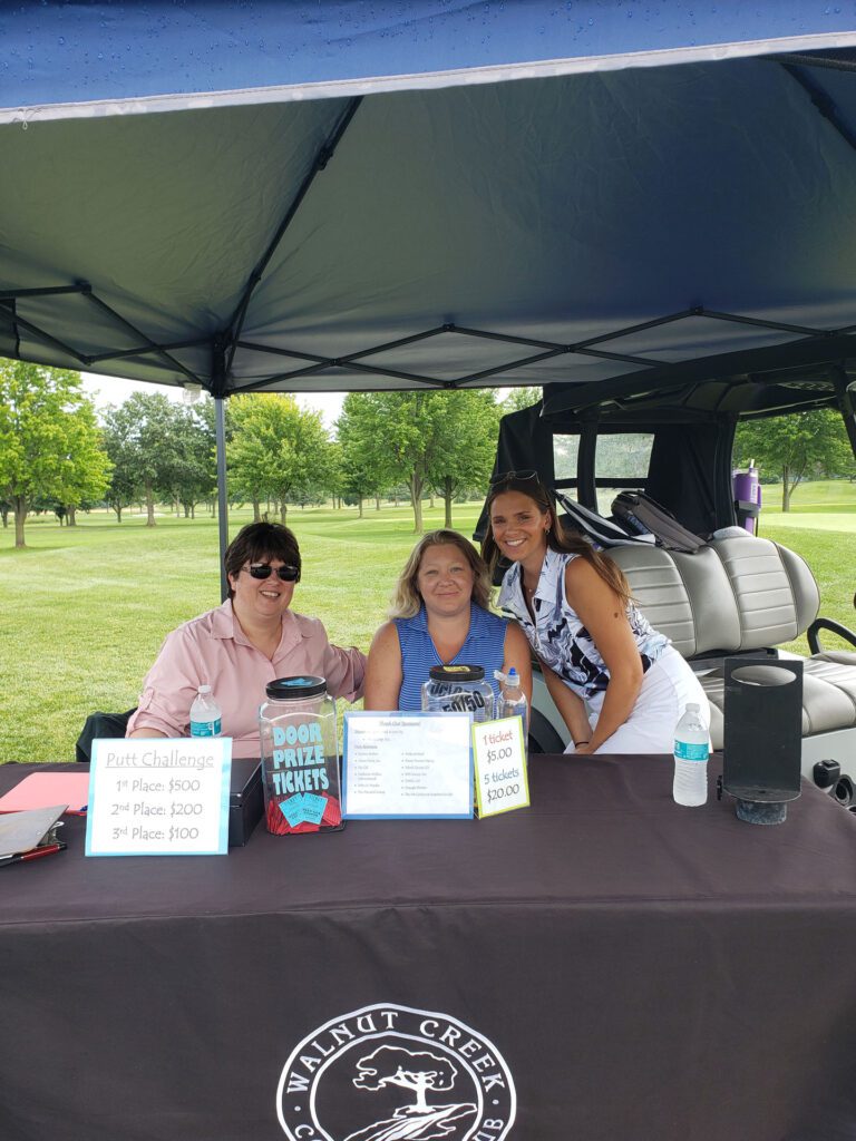 golf-outing-booth Three smiling women seated under a canopy at a Walnut Creek Golf Club table with signs for a putt challenge and door prize tickets, reflecting Sidock Group’s community engagement, teamwork, and philanthropic culture.